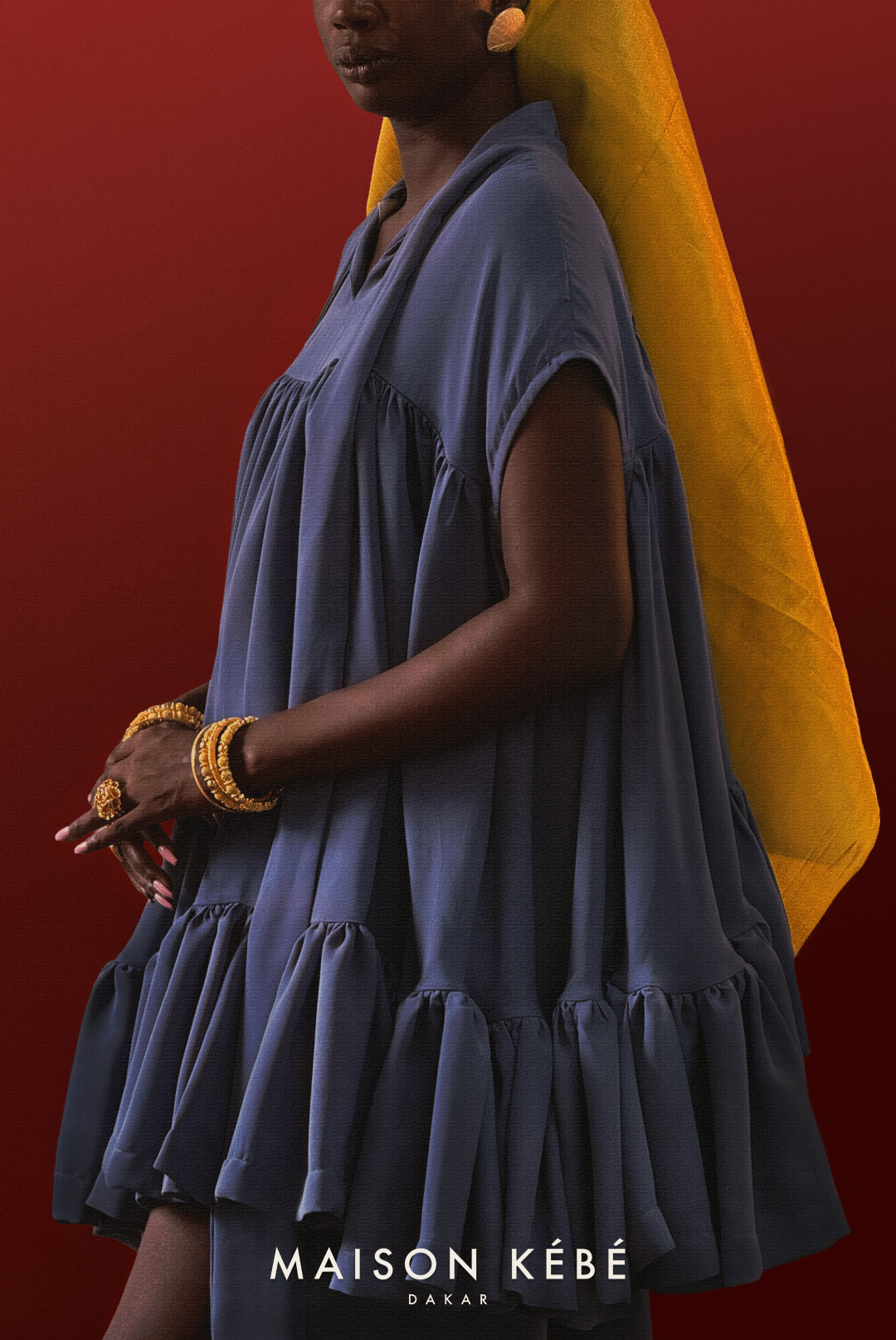 Woman wearing a blue dress with a yellow headscarf against a red background, featuring the Maison Kebe brand.