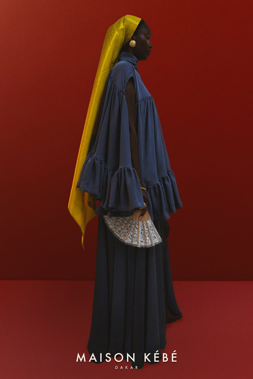 Woman in a blue maison kebe oceane draped outfit with a yellow headscarf against a red background, wearing Maison Kebe.